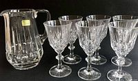 Clear lead crystal pitcher with a ribbed body and angular handle standing beside six matching claret glasses on a black background.