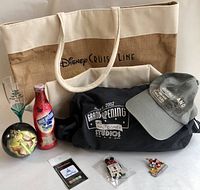 View of the entire lot including Disney Cruise Line tote bag, souvenir glass, commemorative Coca-Cola bottle, ornament ball, souvenir pin, R2D2 USB key, Walt Disney Studios 2002 duffle bag, and Disneyland Paris ball cap.