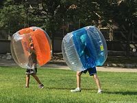 Two people wearing the orange and blue inflatable bumper balls outside on grass.