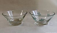 Two small clear glass conical dip bowls side by side on white background, one with turquoise painted leaves and vines, other clear with etched petals.