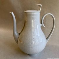 Front view of a white porcelain coffee pot with geometric oval pattern and curved handle and spout.