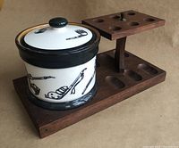 Angled side view of walnut wood pipe stand with ceramic jar placed on base, showing pipe slots and jar lid with black and white pipe illustrations