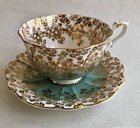 Tea cup and saucer set showing gold filigree and rose motifs on icy blue and white background with scalloped gold rim.