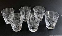 Six short crystal tumblers arranged in two rows on a dark surface, showing their side profiles and etching patterns.