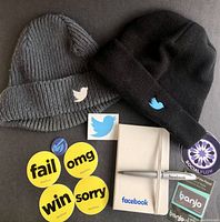 Photo showing two knit toques with Twitter bird logos, Facebook mini notebook with pen, and multiple branded stickers from BuzzFeed, Social Flow, and Banjo.