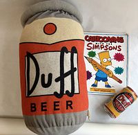 Duff Beer plush pillow, Cartooning with The Simpsons paperback guide, and collectible Duff Beer can arranged together.