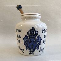 Front view of vintage ceramic Grey Poupon mustard jar showing detailed blue crest graphic and text on creamy white background, with lid and wooden spoon in place.