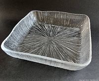 Clear square glass dish with textured starburst pattern radiating from center and wavy edges on black background.