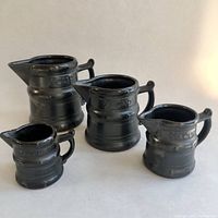 Four black-brown ceramic measuring cups shaped like small pitchers with spouts and handles arranged side by side, showing size differences.