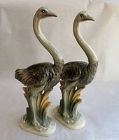 Side view of two ceramic ostrich figurines showing their profile, detailed feathers, and art-deco foliage bases.