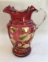 Front and full view of the vintage ruby red glass pitcher with clear handle and gold floral gilding on body and base, showing ruffled top edge.
