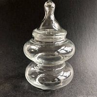 Clear glass apothecary-style stacking bowl with lid shown against black background. Squat, two-tiered rounded shape with bulbous sections and rounded top finial on lid.