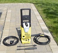 Front view of yellow and black Karcher power washer with coiled GFCI power cord, high-pressure hose and two spray nozzles laid out on patio.