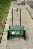 Front view of green plastic drop spreader with large black wheels and handle on paved walkway next to grass.