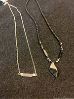 Photo of two necklaces on dark surface. Left necklace has rose quartz rectangular bar pendant on silver chain, right necklace has black onyx kind of beads and a swirl-shaped black onyx pendant.