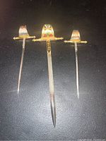 Set of three miniature swords with painted decorative handles and metal blades, laid on black surface.