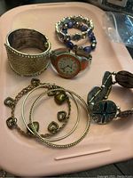 All bracelets and watch arranged on a light pink tray showing various styles and materials including metal, beads, leather, and chain with charms.