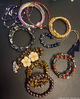 Photo showing 10 assorted bracelets including beaded bracelets, metal bangles, wood bead bracelets, shell flower design, and tassel accented bracelet.