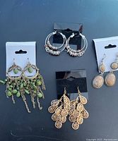 Four pairs of earrings displayed flat on a surface: gold-tone chandelier style with green beads, silver-tone hoop earrings with rings, gold-tone filigree cluster earrings, and gold-tone oval drop earrings with textured detail.