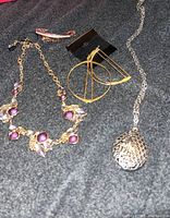 Four jewelry pieces displayed together on dark gray fabric: a gold-tone amethyst necklace, silver-tone egg locket with chain, gold-tone geometric hoop earrings, small rhinestone bar pin.