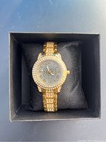 Front view of a gold-tone ladies wristwatch adorned with multiple rhinestones on bezel, dial, and bracelet links, displayed on black cushion inside a black box.