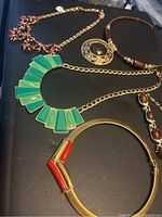 Photo of five antique necklaces spread on a dark surface showing red/gold chain, filigree pendant, brown beads with tassel, green geometric pieces, and gold/red tubular accents.