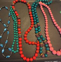Photo showing four beaded necklaces in blue, green, red with multicolored striped beads, and pink beads on a dark surface.
