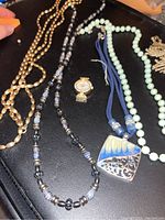  assorted jewelry including 3 beaded necklaces, one gold-tone, one pale green, one black and white, and a small watch pendant on a blue loop