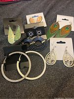 Photo showing seven pairs of earrings with different colors and shapes laid out on a grey fabric surface.