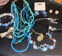 Multiple jewelry pieces laid out on black surface showing various necklaces, bracelets, brooch, charms, and earrings. Colors feature blues, black, coral, and silver-tone metals.