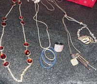 Photo showing long silver-tone necklace with red oval gemstones and other necklaces with hoop and leaf pendants.