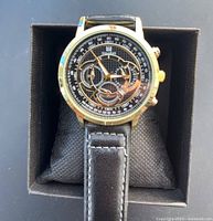 Front view of the new man's Shaarms watch in its display box, showing gold-tone case, black dial with white and gold markings, three sub-dials, and black leather strap with white stitching.