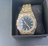 Men's wrist watch shown inside black box with pillow, having black dial with silver markers and ornate gold and black metal band.