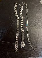 Photo showing three men's bracelets laid out on a dark surface including two thick silver-tone curb chain bracelets and one thinner silver-tone chain bracelet with a plate and blue accent.