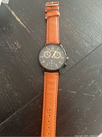 Men's wristwatch laid flat showing black round dial with analog 12-hour display and three subdials, brown leather strap fully extended.