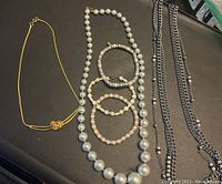 Photo of six jewelry items: various necklaces and bracelets including pearl beads, metallic chains, and gold-tone pieces on a black surface.