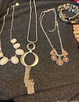 Top-down photo showing three necklaces with silver chains, including a white chunky bead necklace, a silver pendant necklace with tassels, and a coral-colored multi-pendant necklace, beside two bracelets (multicolored beaded and rose-gold cuff).