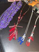 Photo showing four necklaces including one multi-strand purple beaded necklace and three with beaded tassels in red, blue, and mixed colors on chains.