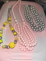 Photo of three necklaces on a pink tray: yellow beaded necklace with silver accents, pink multi-strand beaded necklace, large silver bead choker.