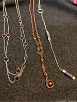Photo shows three distinctive necklaces laid out on a dark fabric background. Each necklace features unique designs with gemstone-like elements in silver, copper, and blue tones.