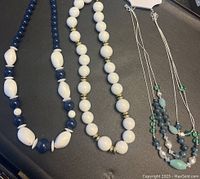 Photo shows three necklaces laid out on a dark surface: two are made of large beads in blue and white shades, and one is a multi-strand silver-tone chain with assorted green, blue, and white beads.