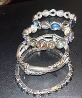 Five silver tone bracelets on a black surface including elastic and metal stretch bands, some with colorful gemstone accents