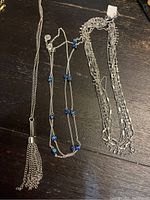 Image showing three silver-tone necklaces laid out on a dark surface. From left to right: a long tassel pendant necklace, a delicate necklace with blue beads, and a multi-strand chain necklace.