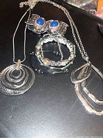 Photo showing several silver-tone necklaces and bracelets arranged on a dark surface including a circular pendant necklace, a blue stone bracelet, and multiple silver textured bangles.