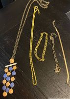 Photo of 5 woman’s jewelry pieces on dark wood surface: 4 gold-tone chains including one with orange and blue circular hanging pendants, 1 wide gold-tone chain bracelet.