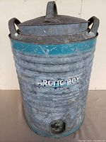 Front view of large antique galvanized steel water cooler showing ribbed body, lid with handle, side handles, and spigot, with visible surface wear.