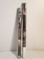Two rolled music posters, one John Lennon and one Kurt Cobain, sealed in original plastic packaging and standing against a plain wall.