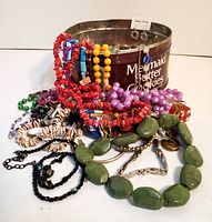 Multiple colorful vintage necklaces including red, orange, green, purple, black beads and silver-tone earrings shown inside a metal tin