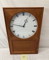 Front view of the wooden 20th century shaker style wall clock with white face and black Roman numerals, closed glass door.