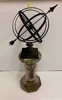 Full view of metal armillary sundial sculpture on weathered concrete pedestal against white wall.